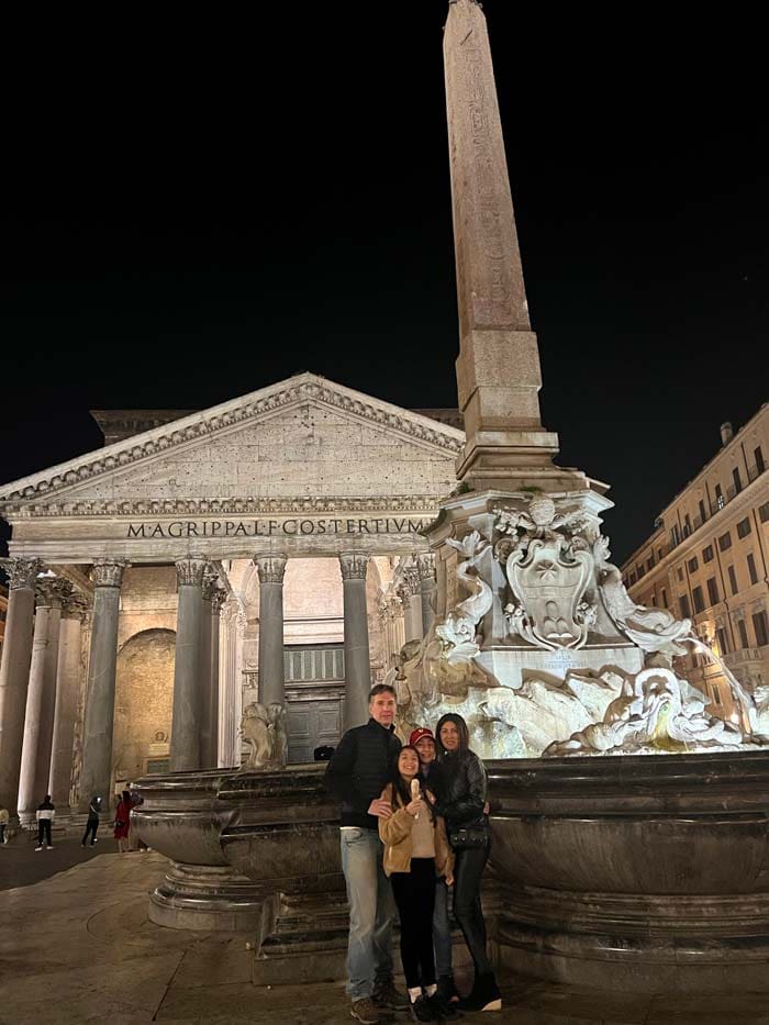 At night, two parents and their two kids stand outside of the Pantheon, a must stop on our Rome itinerary with kids.