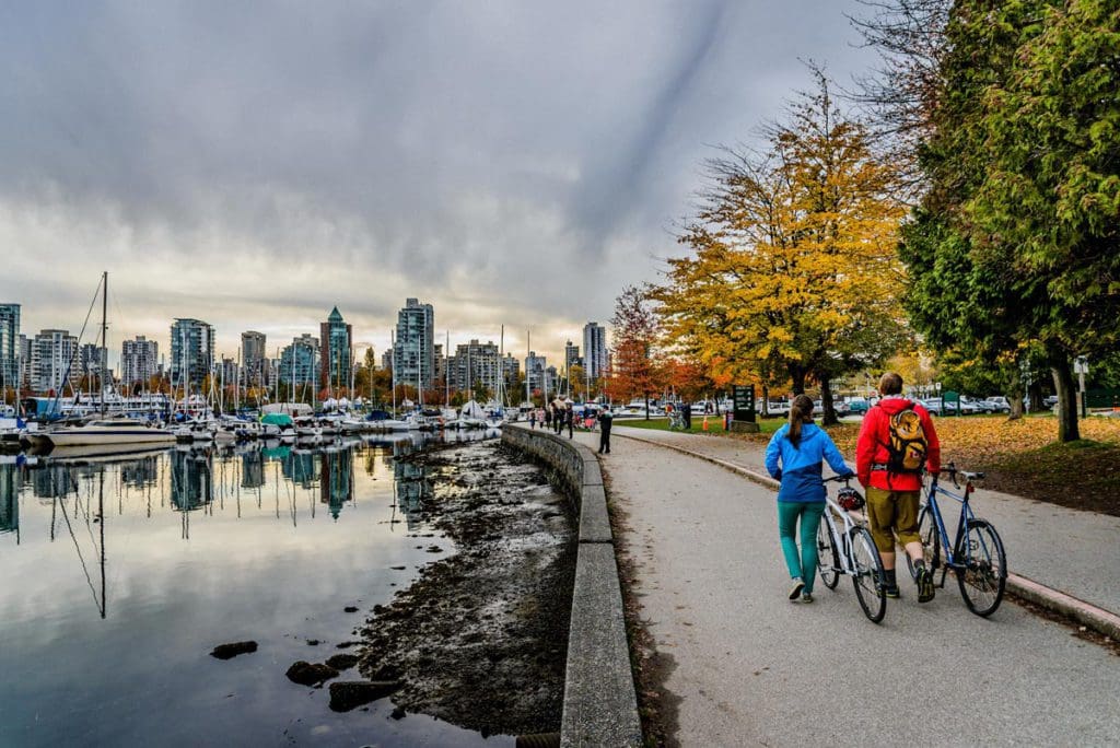 Two people stroll through Stanley Park with the water on their left and the Vancouver skyline in front of them.