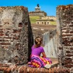 A young girl sits along a brick wall looking at San Felipe del Morro Castle in San Juan.