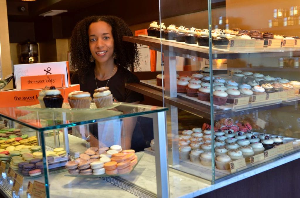 A woman stands behind the corner at The Sweet Lobby, serving several variety of colorful cupcakes.
