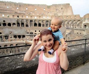A mom holds her infant son on her shoulders, while walking around the Colosseum in Rome.
