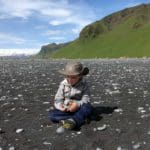 A young boy with an explorer hat sits along the black beach in Iceland with green hills in the distance.