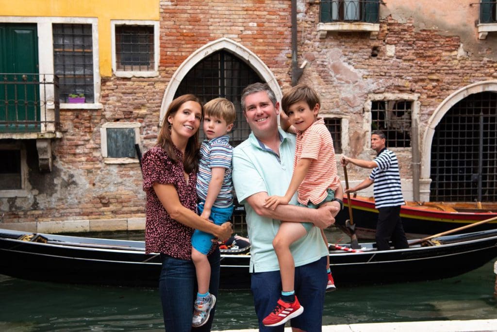 A family of four stands together with a Venetian canal and gondola behind them. 