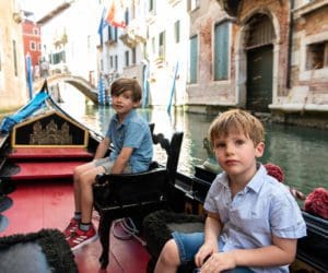 Two young boys ride in a gondola down a canal in Venice, Venetian buildings behind them.
