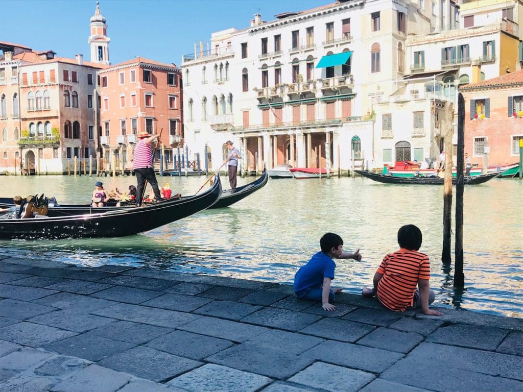 Two kids sit alongside a canal in Venice, while gondolas pass by on a sunny day.