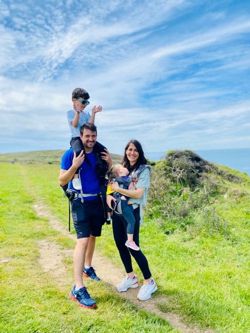 A family of four stands together in the Welsh countryside with the sea behind them, dad is holding a small boy on his shoulders, while mom is wearing a smaller child on her chest.