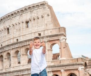 A young boy in front of the Colosseum in Rome, Italy, giving a thumbs up with both hands.