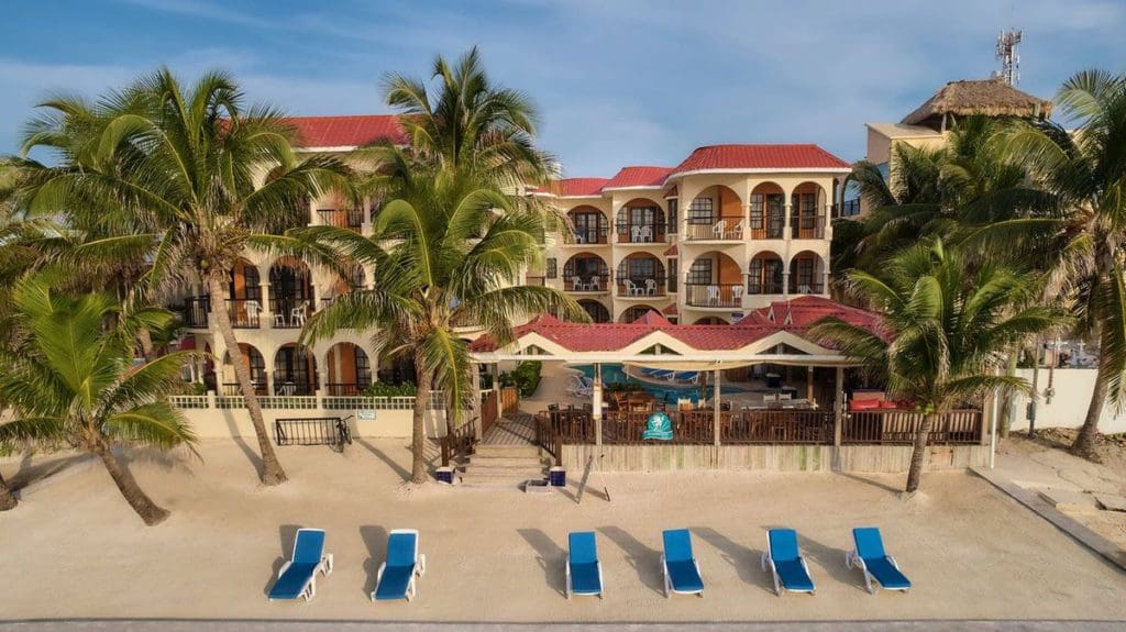Six blue beachside loungers sit on the beach with the resort buildings for Sunbreeze Belize in the distance.