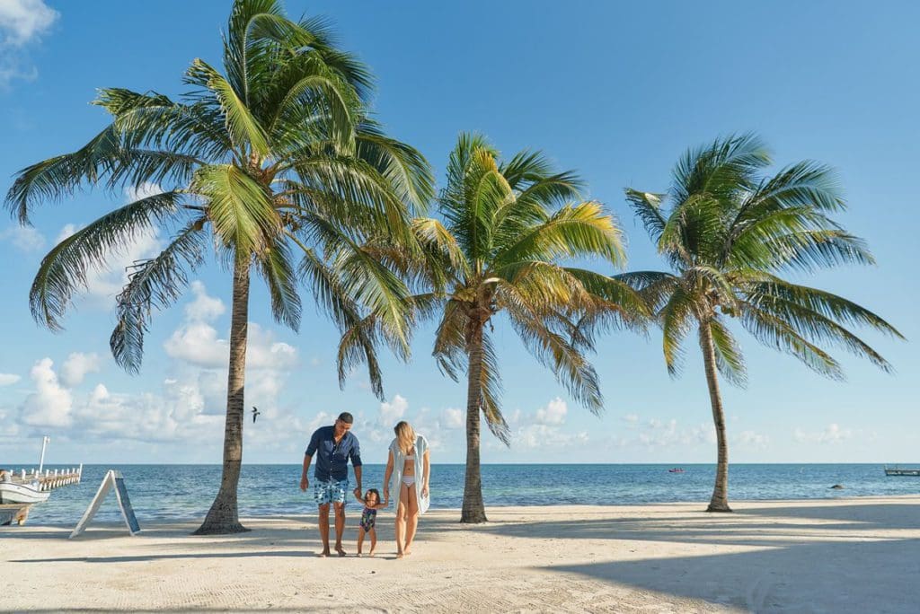 A young couple walks their infant along the beach with swaying palm trees overhead at Photo Courtesy: Sunbreeze Suites in Belize.
