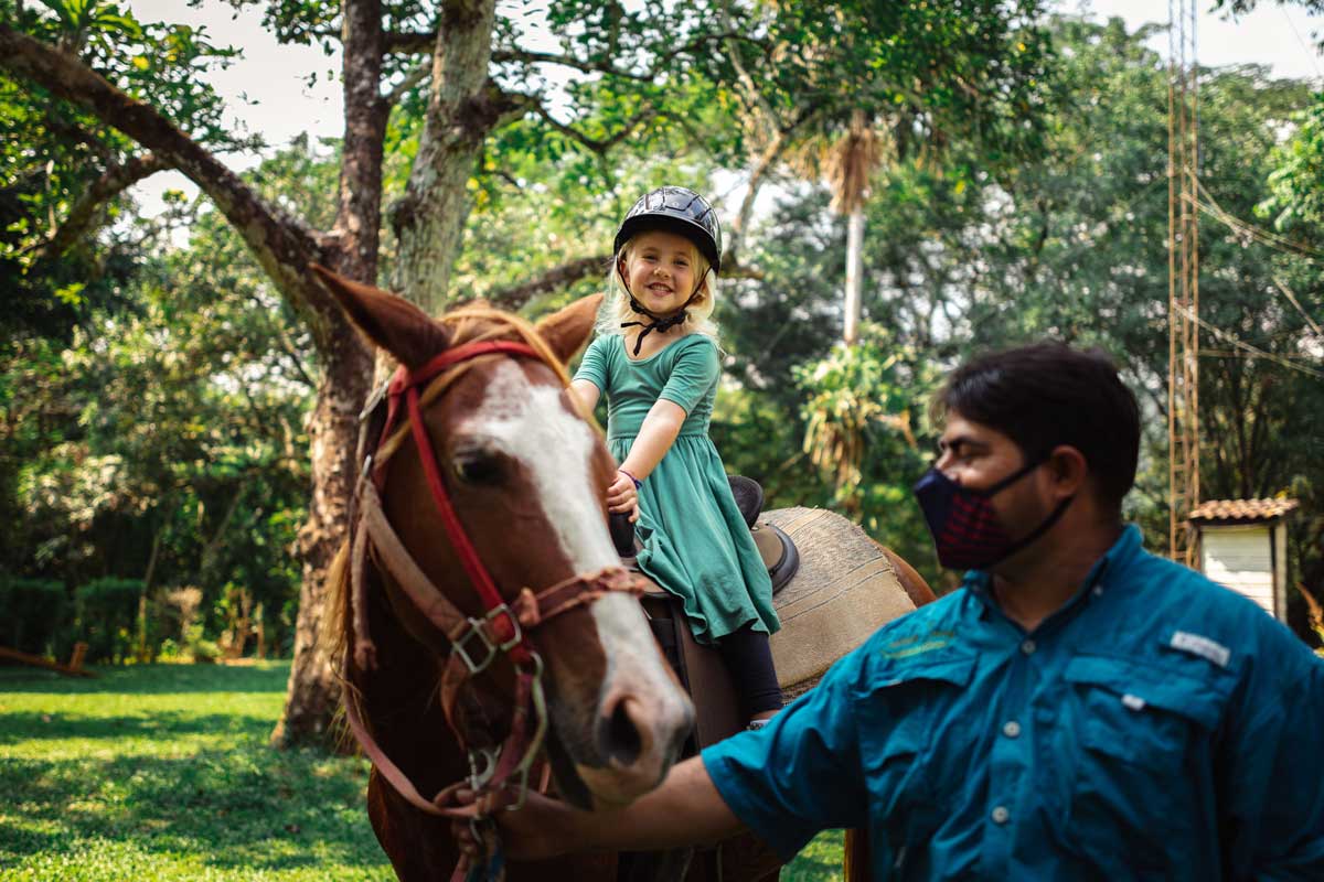 A young girl sits on a horse, while a staff member leads it along a path at Sweet Songs Jungle Lodge, a Muy’Ono Resort.