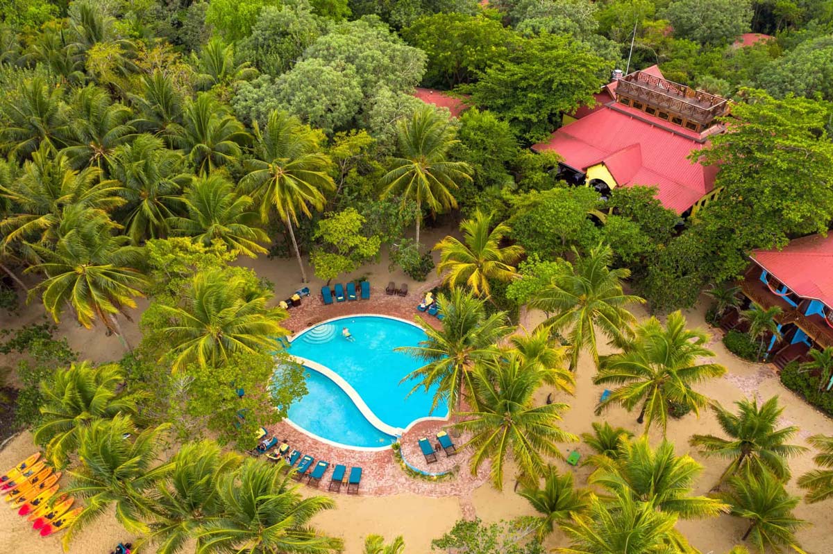 An aerial view of the intimate pool at Hamanasi Adventure & Dive Resort, with red-roofed resort buildings on one side and lush foliage around the grounds.