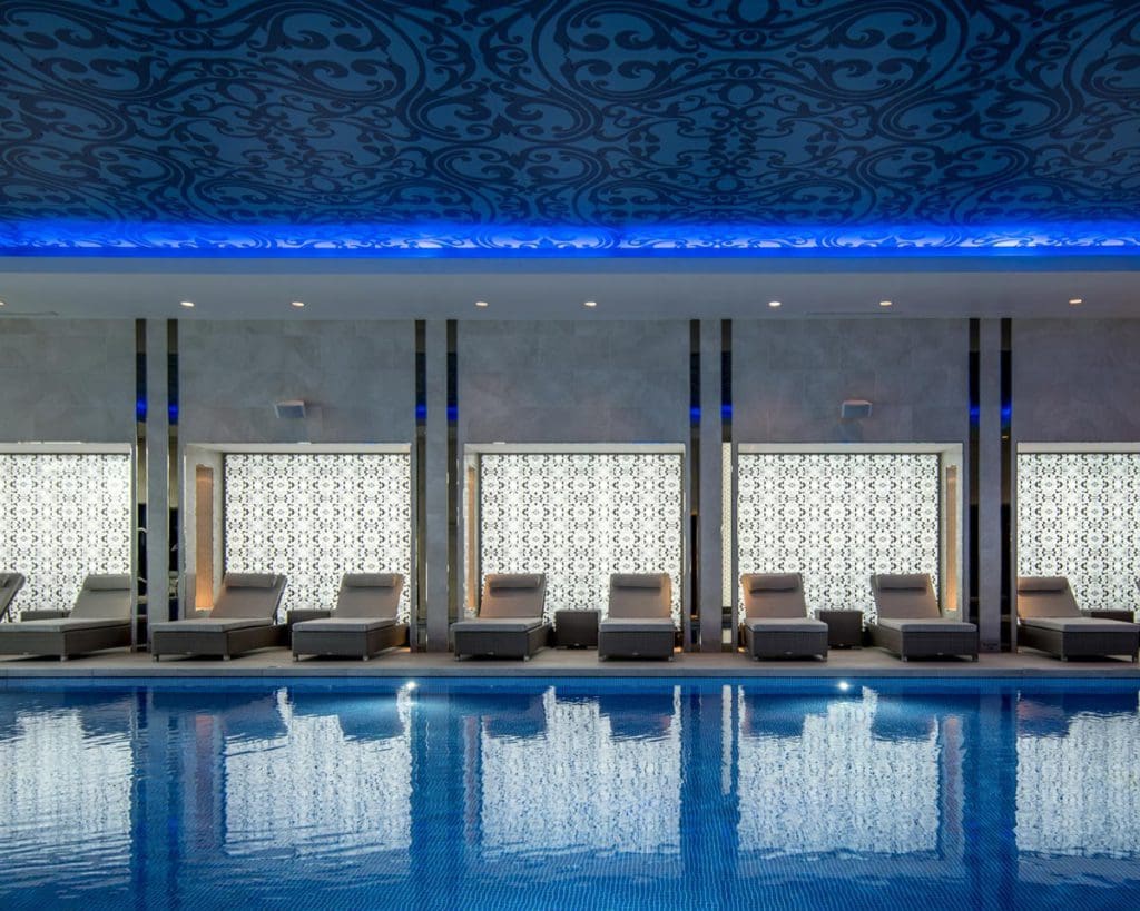 The blue-hued pool area of Intercontinental London - The O2, An IHG Hotel, featuring a pool and loungers flanking the pool on one side.