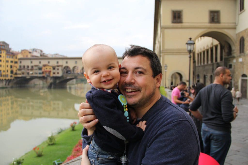 A dad holds a young baby with Ponte Vecchio in the distance.