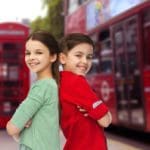 Brother and sister stand back to back, smiling, with iconic London imagery behind them, including a red phone booth and double decker bus.