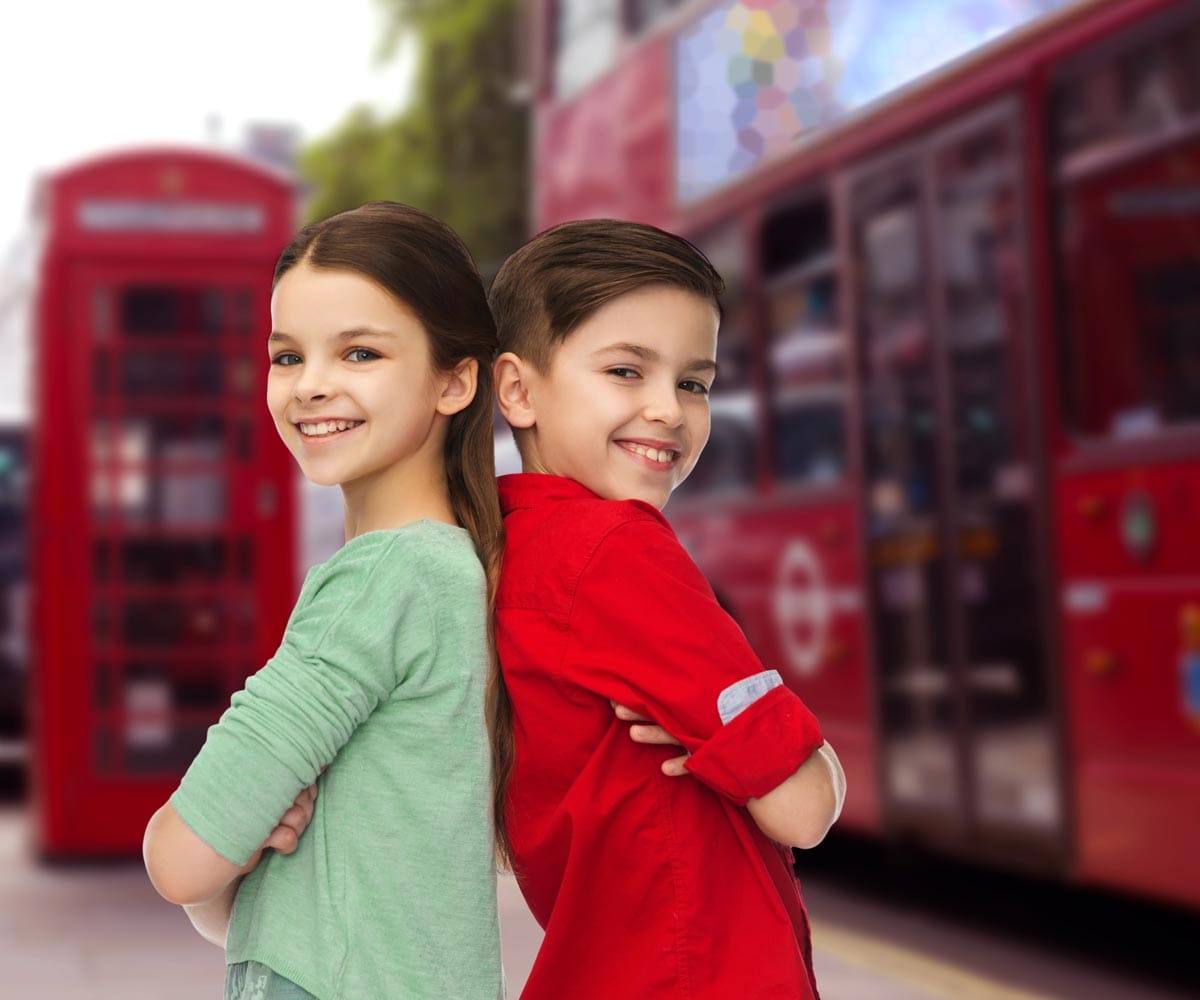 Brother and sister stand back to back, smiling, with iconic London imagery behind them, including a red phone booth and double decker bus.