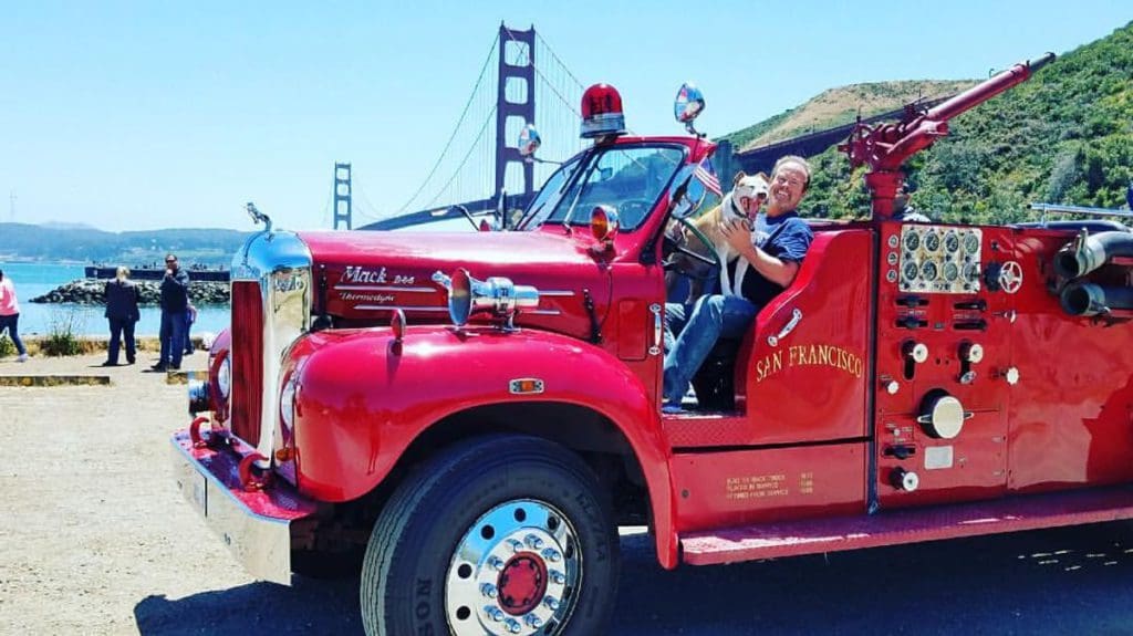 A man sits on a firetruck with a dog, with the Golden Gate Bridge in the distance.
