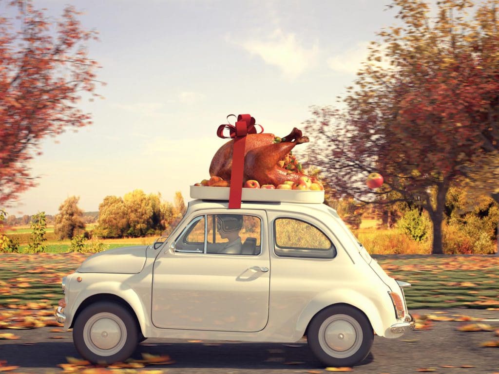 A small car drives through a fall landscape with a huge cooked turkey strapped to the roof.