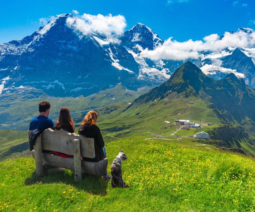 A family of three sits together on a bench, while taking in a mountain view near Wengen, one of the best towns and villages to visit with your family in Switzerland.