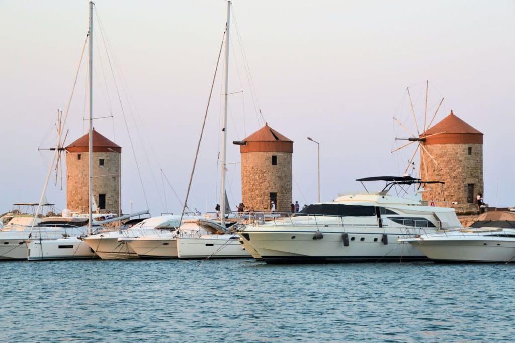 A line of yachts float in the water in front of three iconic windmills on the shore of Rhodes.