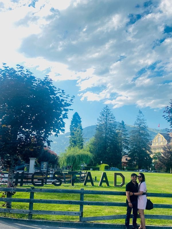 A husband and wife stand together next to a huge sign reading "#GSTAAD" with a pleasant Swiss scene behind them on a sunny day.