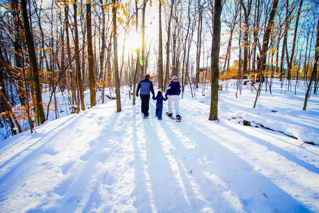 A mom and dad hold the hands of a young child, all on snowshoes through the snow on a trail in Blandford Nature Center near Grand Rapids.