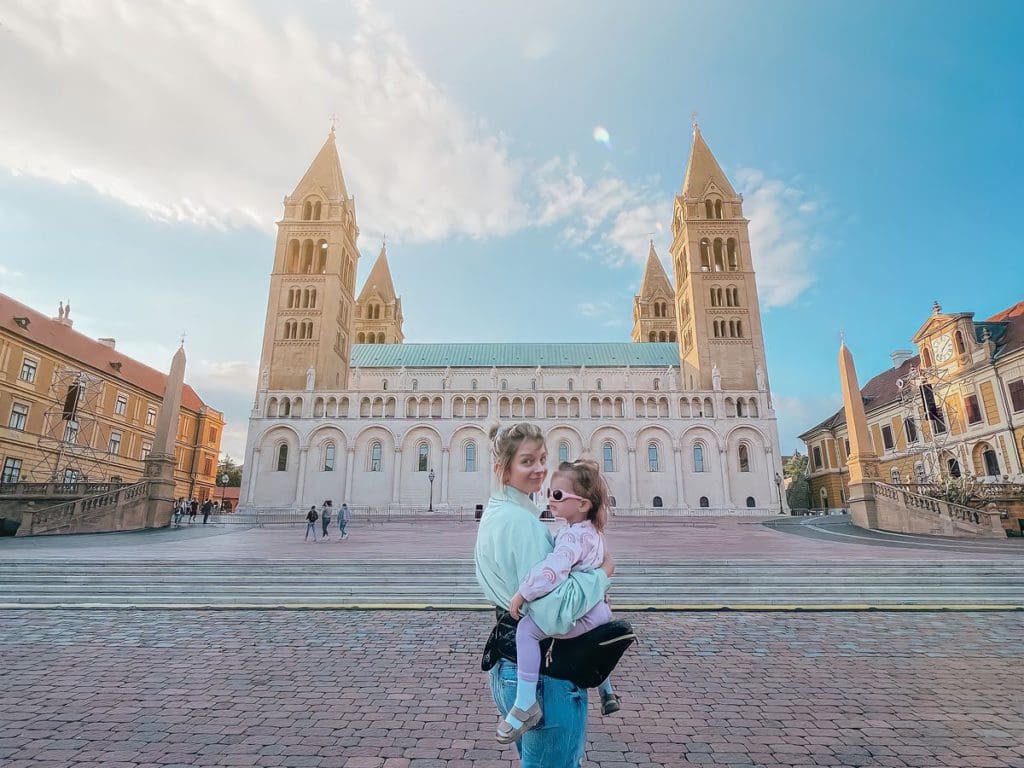 A mom holds her daughter using a hip-hugger to support her toddler, while exploring a European city - a hip-hugger is a must-pack for a European vacation with toddlers.