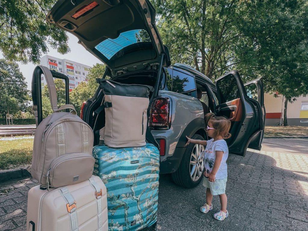 A young girl points at luggage waiting to be packed for a road trip in front of the car.