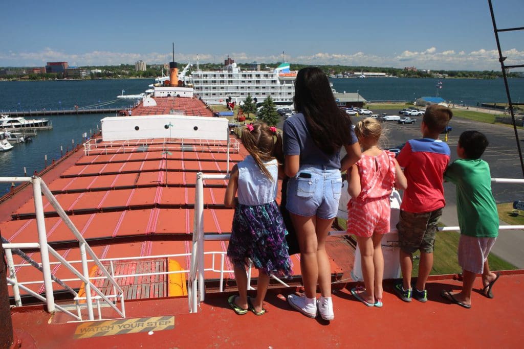 Five kids stand along a ship deck looking out onto the large shipping container ship at Museum Ship Valley Camp in Upper Michigan.