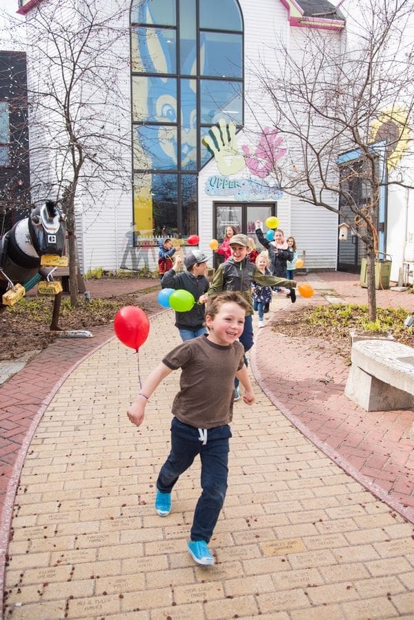 A line of children, all holding colorful balloons, run out of the exit of The Upper Peninsula Children's Museum.