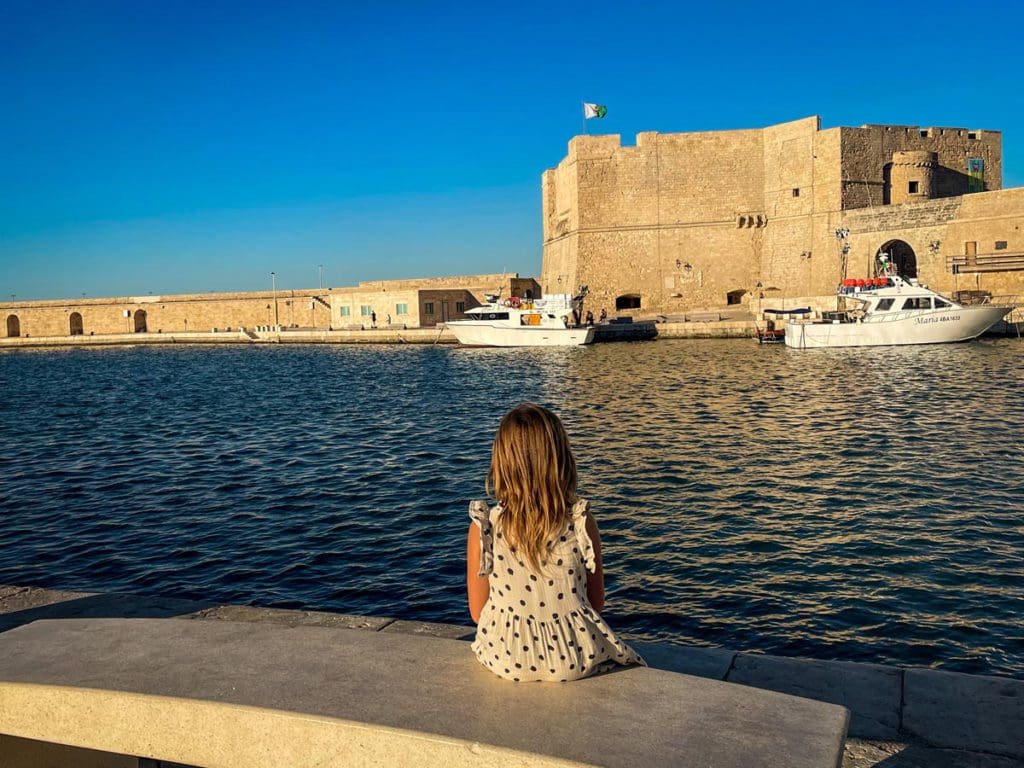 A young girl looks out over a small bay toward a castle of sorts in Monopoli, one of the best places to visit in Italy with kids.