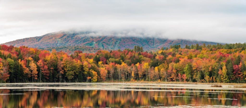 A vibrant view of fall colors in all autumn hues across a lake, with Stratton Mountain Resort in the distance.