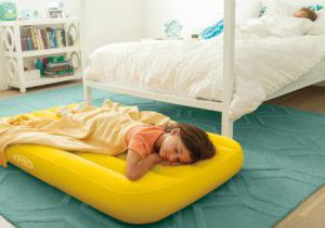 A young girl lays down on a yellow Intex Kids Travel Bed in her room.